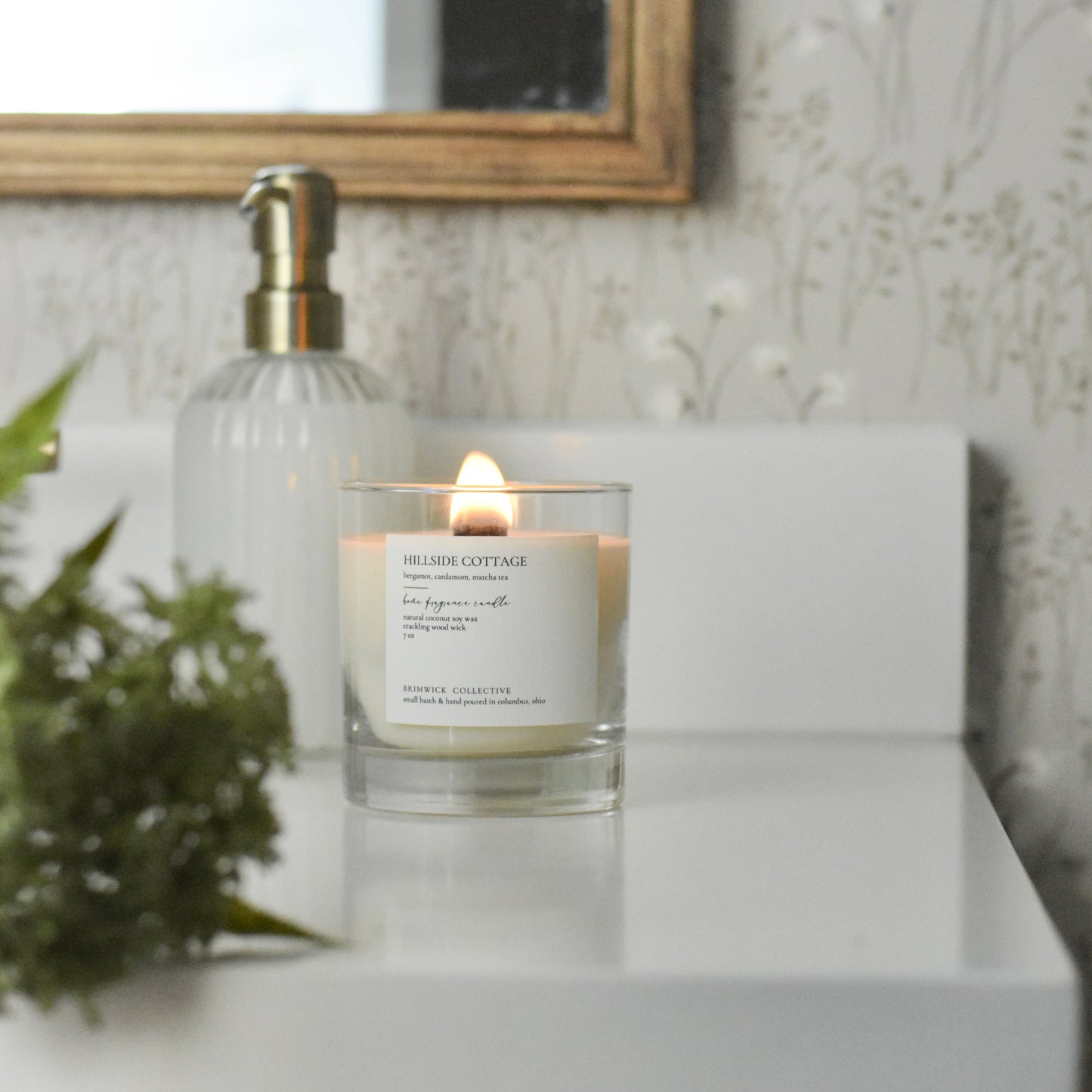 Candle with a visible brand label on a white surface, with a decorative wall and plant in the background.